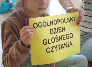 Dziewczynka trzyma kartkę z napisem Ogólnopolski Dzień Głośnego Czytania.