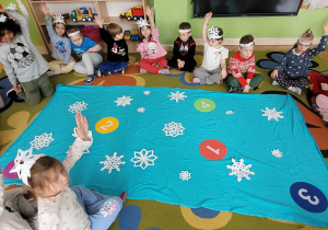 Dzieci siedzą w kole i zgłaszają się do zadania matematycznego, które ma polegać na porządkowaniu gwiazdek śniegowych.