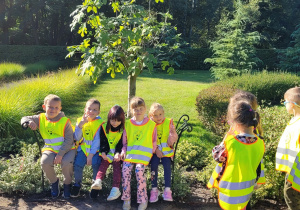 Piątka dzieci siedzi na ławeczce w Ogrodzie Botanicznym, a pozostałe im się przyglądają.