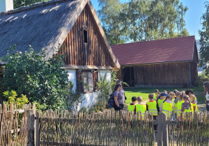 Dzieci słuchają opowieści przewodnika przed skansenem w Ogrodzie Botanicznym.
