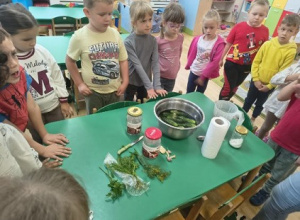 Dzieci stoją wokół stołu, na którym znajdują się produkty do kiszenia ogórków.