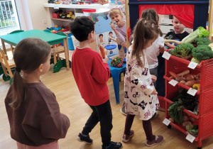 Dzieci stoją w kolejce do straganu warzywnego w klasie, trzymając w ręce warzywa.